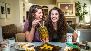 Two women sit at a dining table laughing as one sips from a pineapple with straws held between them in a bright kitchen/living room.