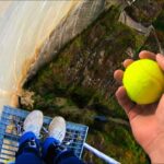 Person wearing blue pants and white sneakers stands on a metal platform high above a canyon, holding a bright yellow tennis ball at arm's length.