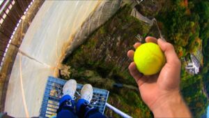 Person wearing blue pants and white sneakers stands on a metal platform high above a canyon, holding a bright yellow tennis ball at arm's length.