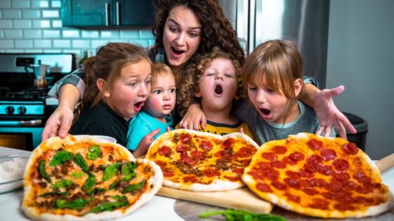 A mother and four children hover over three pepperoni and veggie pizzas on a kitchen counter, mouths agape in excitement.
