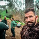 Bearded man in a brown fur cloak takes a selfie on an outdoor film set with crew and equipment in the background; a boom mic hangs overhead.