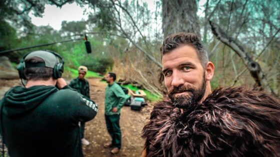 Bearded man in a brown fur cloak takes a selfie on an outdoor film set with crew and equipment in the background; a boom mic hangs overhead.
