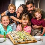 A family of six gathers in a kitchen around a large sushi platter on the counter, smiling at the camera.