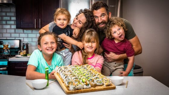 A family of six gathers in a kitchen around a large sushi platter on the counter, smiling at the camera.