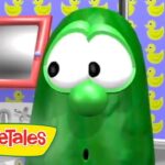Green cucumber character from VeggieTales in a colorful room with yellow rubber-duck wallpaper and a pink-framed window behind him