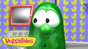 Green cucumber character from VeggieTales in a colorful room with yellow rubber-duck wallpaper and a pink-framed window behind him