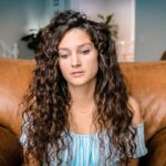 Young woman with long curly brown hair sits on a brown leather couch, looking contemplative in a living room.