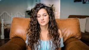 Young woman with long curly brown hair sits on a brown leather couch, looking contemplative in a living room.