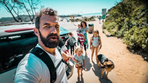 Man with a backpack taking a selfie as his family and their dog walk toward the beach beside a parked SUV.