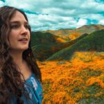 Woman with long dark hair in a blue off-shoulder top stands in a field of orange wildflowers with rolling green hills and a blue sky behind her.
