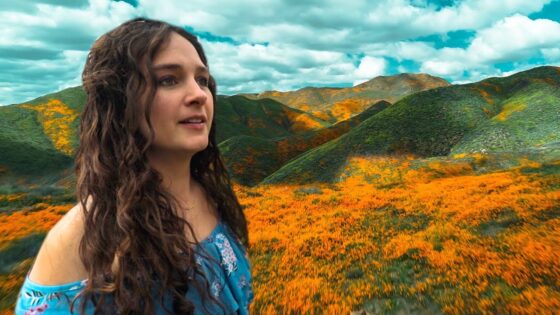 Woman with long dark hair in a blue off-shoulder top stands in a field of orange wildflowers with rolling green hills and a blue sky behind her.