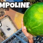 Person standing on a high metal platform, holding a large green watermelon, aerial ground view below (thumbnail for a trampoline video).