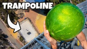 Person standing on a high metal platform, holding a large green watermelon, aerial ground view below (thumbnail for a trampoline video).