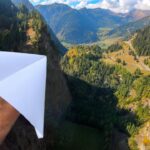 A hand holding a white paper airplane over a steep mountain valley with trees and winding roads below.
