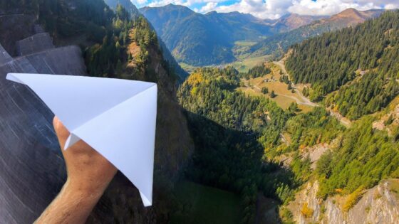 A hand holding a white paper airplane over a steep mountain valley with trees and winding roads below.