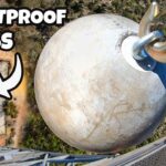 Drone view of a large round ballast sphere attached to a crane on a high-rise balcony, with 'BULLETPROOF GLASS' text overlay