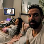 Pregnant woman on an exam table as a nurse uses an ultrasound machine; her partner takes a selfie in the foreground.