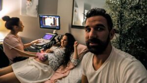 Pregnant woman on an exam table as a nurse uses an ultrasound machine; her partner takes a selfie in the foreground.