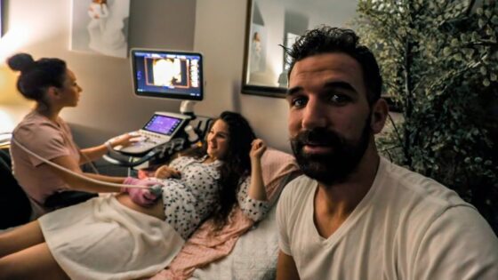 Pregnant woman on an exam table as a nurse uses an ultrasound machine; her partner takes a selfie in the foreground.