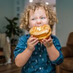 Curly-haired child in a blue shirt bites into a glazed doughnut indoors at home.