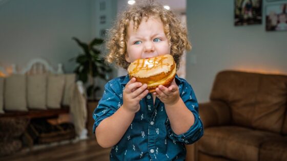 Curly-haired child in a blue shirt bites into a glazed doughnut indoors at home.
