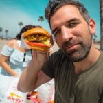 Smiling man on a sunny beach holds a burger toward the camera, with palm trees and a woman in a striped dress in the background.