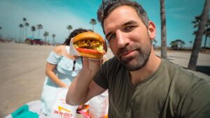 Smiling man on a sunny beach holds a burger toward the camera, with palm trees and a woman in a striped dress in the background.