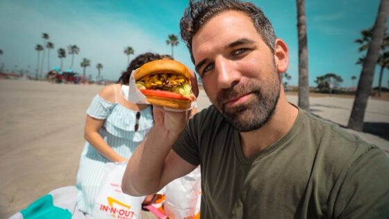 Smiling man on a sunny beach holds a burger toward the camera, with palm trees and a woman in a striped dress in the background.