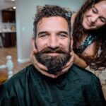 Man with a thick beard wearing a barber cape smiles as a woman behind him gently holds his cheeks.