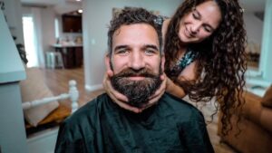 Man with a thick beard wearing a barber cape smiles as a woman behind him gently holds his cheeks.