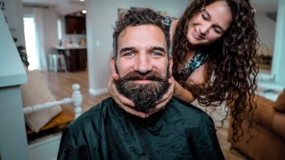 Man with a thick beard wearing a barber cape smiles as a woman behind him gently holds his cheeks.
