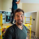 Man in foreground taking a selfie on a yellow construction scaffold inside a room under renovation; smiling wide at the camera.