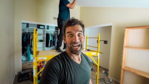 Man in foreground taking a selfie on a yellow construction scaffold inside a room under renovation; smiling wide at the camera.