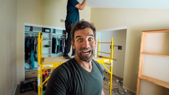 Man in foreground taking a selfie on a yellow construction scaffold inside a room under renovation; smiling wide at the camera.