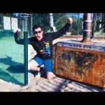 Man in sunglasses with a black graphic shirt kneels in sand at a playground, posing beside a large rusted industrial cabinet and a glass panel glass pane.