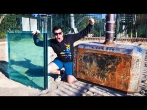 Man in sunglasses with a black graphic shirt kneels in sand at a playground, posing beside a large rusted industrial cabinet and a glass panel glass pane.