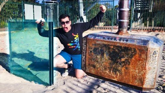 Man in sunglasses with a black graphic shirt kneels in sand at a playground, posing beside a large rusted industrial cabinet and a glass panel glass pane.