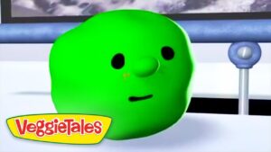 Close-up of a bright green cucumber character with black eyes and a small mouth, from VeggieTales, resting on a white surface with a logo in the corner.