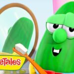 A green VeggieTales cucumber character smiles while brushing teeth with an orange toothbrush in front of a round mirror in a bright scene.