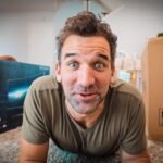 Man lying on a carpeted floor between boxed items, looking up with a surprised expression toward the camera (moving day). My left: an LG TV box; right: a large cardboard box.