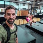 Man wearing a harness takes a selfie in an indoor obstacle-course gym; two kids swing on ropes in the background.