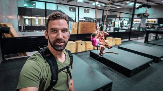 Man wearing a harness takes a selfie in an indoor obstacle-course gym; two kids swing on ropes in the background.