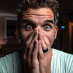 Man with wide eyes covering his mouth with both hands, surprised expression in a dimly lit room