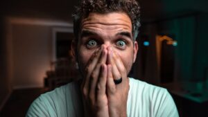 Man with wide eyes covering his mouth with both hands, surprised expression in a dimly lit room