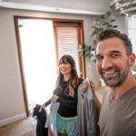 Man in a gray shirt takes a selfie in a bright living room as a woman holds clothing and a photographer records the moment in the background.