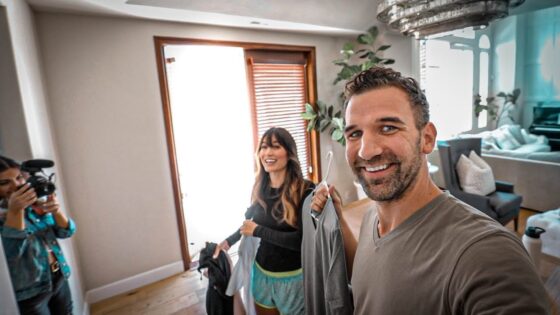 Man in a gray shirt takes a selfie in a bright living room as a woman holds clothing and a photographer records the moment in the background.