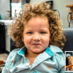 Child with curly hair sits in a barber chair, smiling at the camera in a salon setting