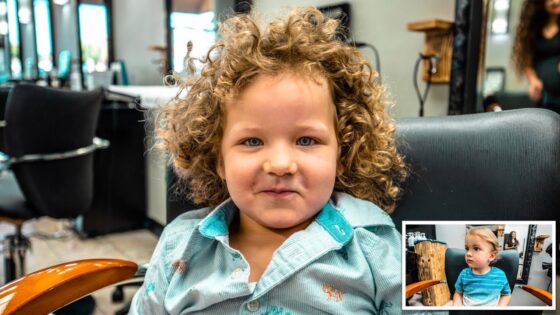 Child with curly hair sits in a barber chair, smiling at the camera in a salon setting