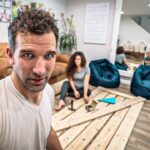 Man takes a selfie in a living room with a woman sitting on the floor among wooden planks and tools for a project.
