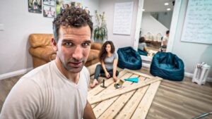 Man takes a selfie in a living room with a woman sitting on the floor among wooden planks and tools for a project.